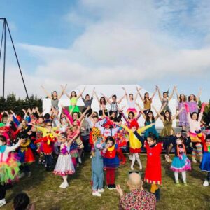 Groupe d'enfants et d'adultes en costumes colorés, bras levés, participant à un spectacle en plein air sur une scène avec un ciel dégagé en arrière-plan.