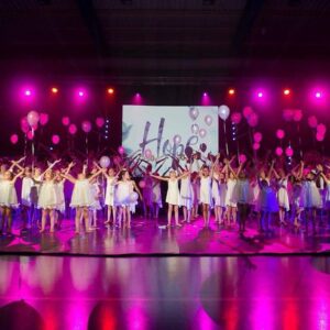 Jeunes danseuses en robes blanches tenant des ballons roses, sur une scène illuminée par des lumières violettes, avec le mot 'Hope' affiché en arrière-plan.
